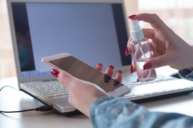 Jovem mulher com máscara facial desinfecção de superfícies de gadgets no seu local de trabalho