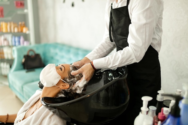 Jovem mulher com máscara facial protetora desfrutando durante a lavagem do cabelo no cabeleireiro
