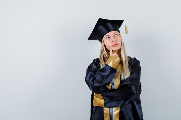 Jovem mulher com o dedo no queixo em uniforme de pós-graduação e parecendo pensativa