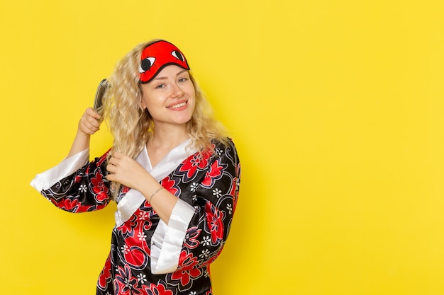 Jovem mulher com roupão de noite e máscara de olho escovando o cabelo de frente, sorrindo na parede amarela modelo de cama noturna para menina