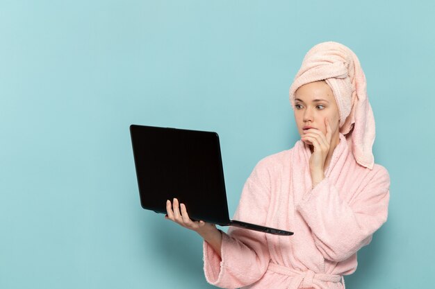 jovem mulher com roupão rosa, depois do banho, vendo algo no laptop na mesa azul