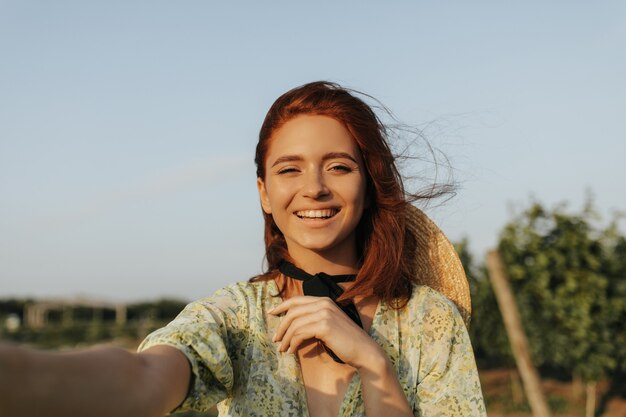 Jovem mulher com sardas, cabelo ruivo e bandagem preta no pescoço em roupas verdes estampadas sorrindo e tirando foto ao ar livre