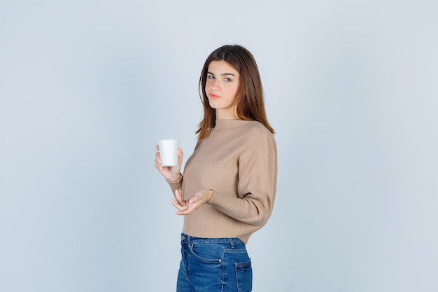Jovem mulher com suéter, jeans mantendo o copo de papel, ficando descontente com a pergunta idiota e parecendo melancólica.