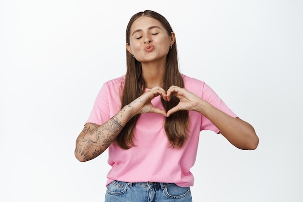 Foto grátis jovem mulher com tatuagens franzindo os lábios para beijo, mostra o sinal da mão do coração e fecha os olhos, de pé bobo contra o fundo branco