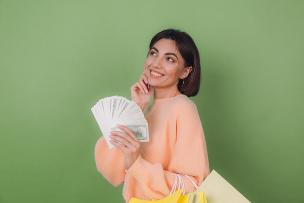 Jovem mulher com um suéter casual cor de pêssego isolado em uma parede verde-oliva segurando um leque de notas de 100 dólares, dinheiro e sacolas de compras, pensando positivo, sorrindo, espaço de cópia