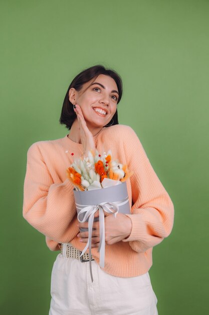 Jovem mulher com um suéter casual cor de pêssego isolado na parede verde-oliva segura uma caixa de flores brancas de laranja com flores de algodão, gypsophila, trigo e lagurus, para um presente feliz espantado surpreso