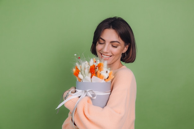 Foto grátis jovem mulher com um suéter casual cor de pêssego isolado na parede verde-oliva segura uma caixa de flores brancas de laranja com flores de algodão, gypsophila, trigo e lagurus, para um presente feliz espantado surpreso