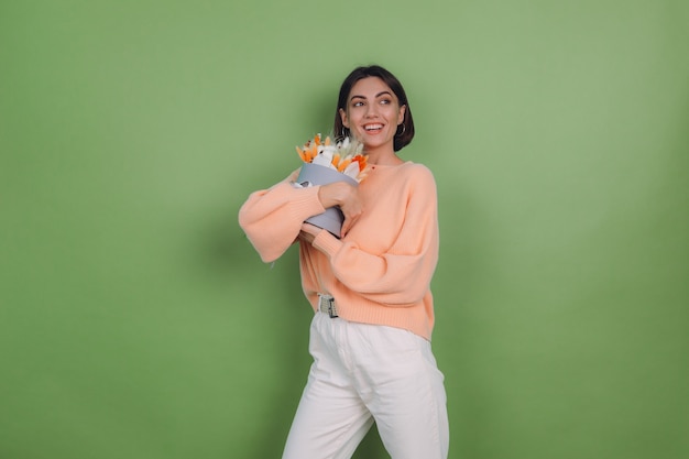 Foto grátis jovem mulher com um suéter casual cor de pêssego isolado na parede verde-oliva segura uma caixa de flores brancas de laranja com flores de algodão, gypsophila, trigo e lagurus, para um presente feliz espantado surpreso