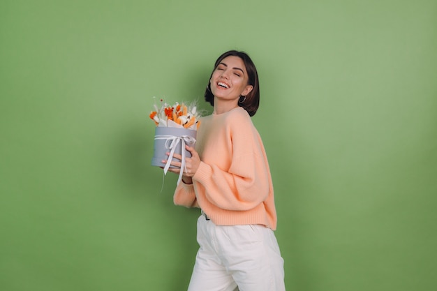 Jovem mulher com um suéter casual cor de pêssego isolado na parede verde-oliva segura uma caixa de flores brancas de laranja com flores de algodão, gypsophila, trigo e lagurus, para um presente feliz espantado surpreso