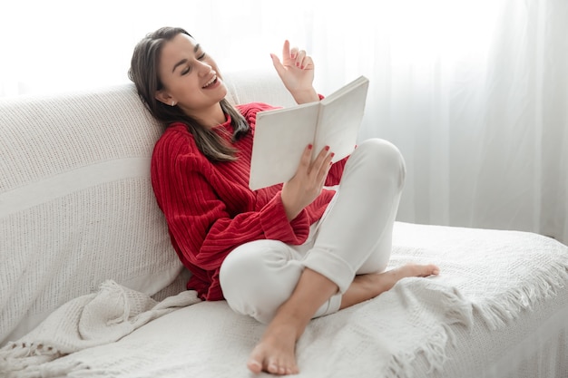 Jovem mulher com um suéter vermelho no sofá em casa com um livro nas mãos.