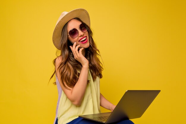 Jovem mulher com um vestido amarelo com chapéu e óculos escuros, ouvindo música, falando ao telefone e usando o laptop