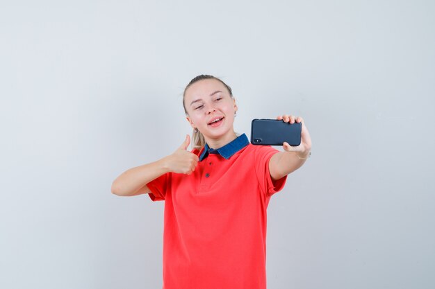 Jovem mulher com uma camiseta mostrando o polegar para cima enquanto tira uma selfie e parece alegre