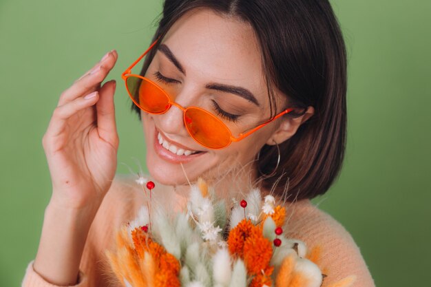 Jovem mulher com uma camisola pêssego casual isolada na parede verde-oliva segura uma composição de caixa de flores brancas e laranjas de flores de algodão, gipsófila, trigo e lagurus para um presente feliz surpresa