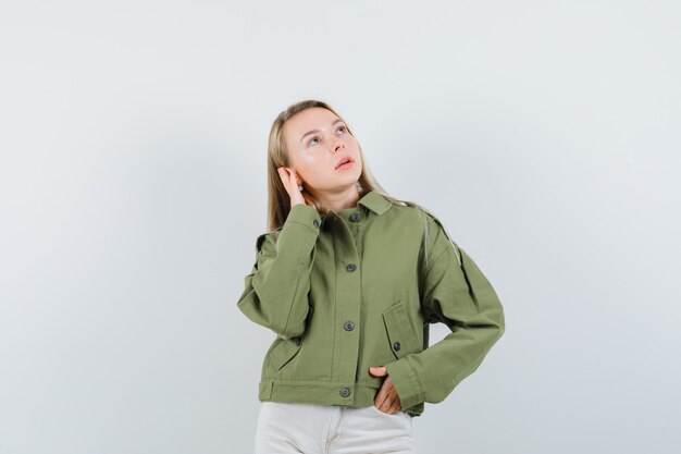 Jovem mulher com uma jaqueta verde, jeans posando com a mão na orelha e olhando impressionante, vista frontal.