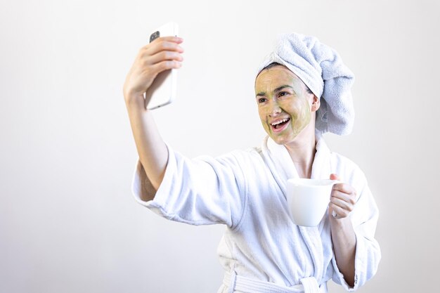 Jovem mulher com uma máscara de beleza verde no rosto com uma xícara e um smartphone