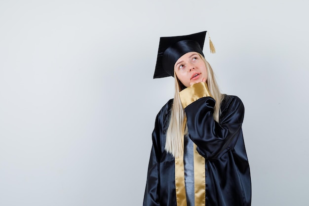 Jovem mulher com uniforme de pós-graduação, segurando o queixo e parecendo pensativa