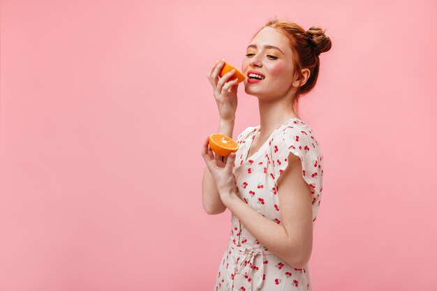 Jovem mulher com vestido de verão branco gosta de laranja doce em fundo rosa.