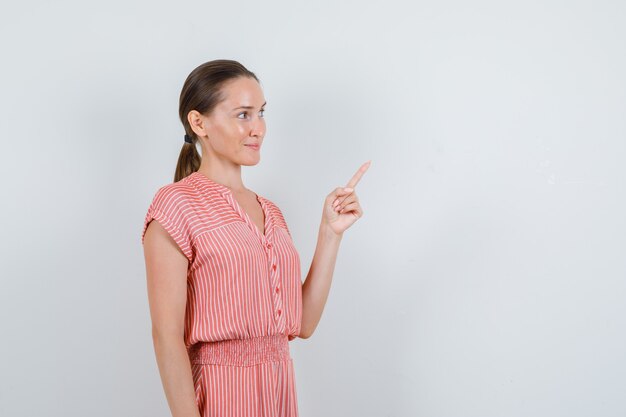 Jovem mulher com vestido listrado, apontando para o lado e olhando alegre, vista frontal.