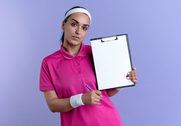 Jovem mulher confiante e desportiva, caucasiana, usando bandana e pulseiras, segurando uma caneta e uma prancheta isoladas no espaço roxo com espaço de cópia