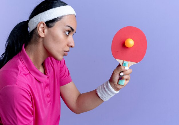 Jovem mulher confiante e desportiva, caucasiana, usando bandana e pulseiras, segurando uma raquete de pingue-pongue e uma bola isolada no espaço roxo com espaço de cópia