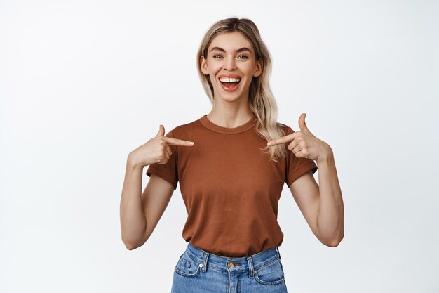 Jovem mulher confiante sorrindo e apontando os dedos para si mesma mostrando na camiseta em pé contra o fundo branco