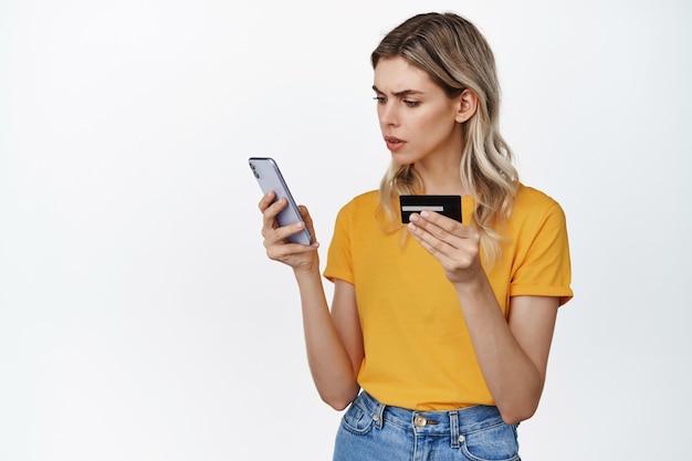 Foto grátis jovem mulher confusa, segurando o cartão de crédito e olhando para a tela do smartphone com cara de perplexa, conceito de problema com transferência de dinheiro ou pagamento em app em branco.