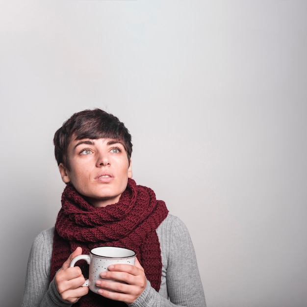 Jovem mulher contemplada que guarda a caneca de café branca contra o fundo cinzento