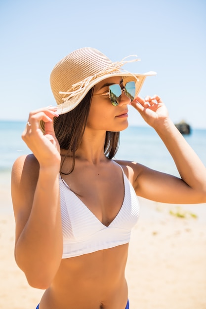 Jovem mulher de biquíni com chapéu de palha branco, aproveitando as férias de verão na praia. Retrato de uma linda mulher latina relaxante na praia com óculos de sol.