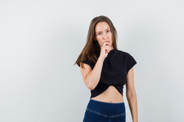 Jovem mulher de blusa preta, calça segurando a mão no queixo e parecendo hesitante