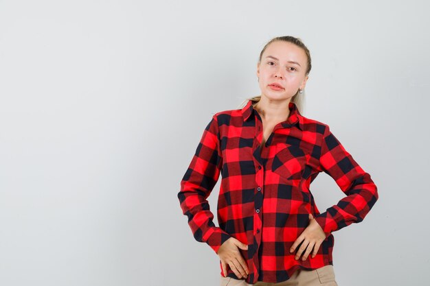 Jovem mulher de camisa xadrez, calças posando em pé e com uma bela vista frontal.