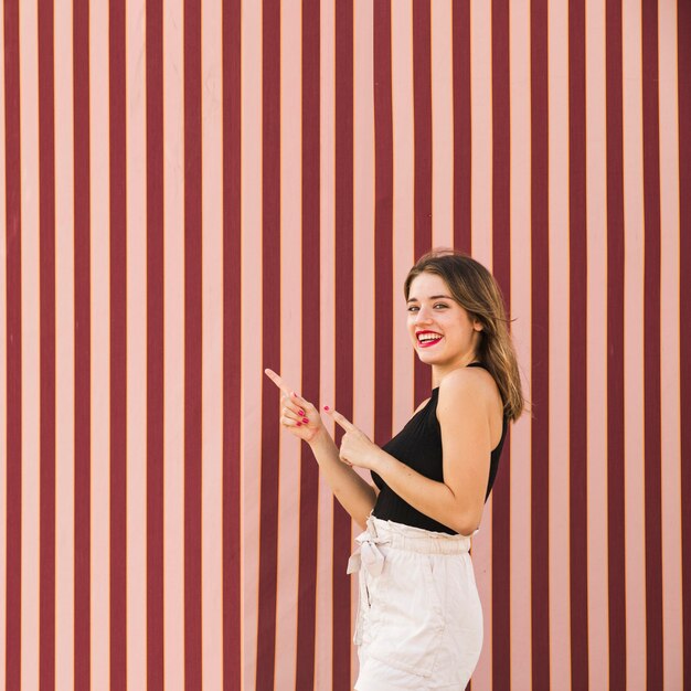 Jovem mulher de sorriso que está na frente do contexto listrado que aponta os dedos