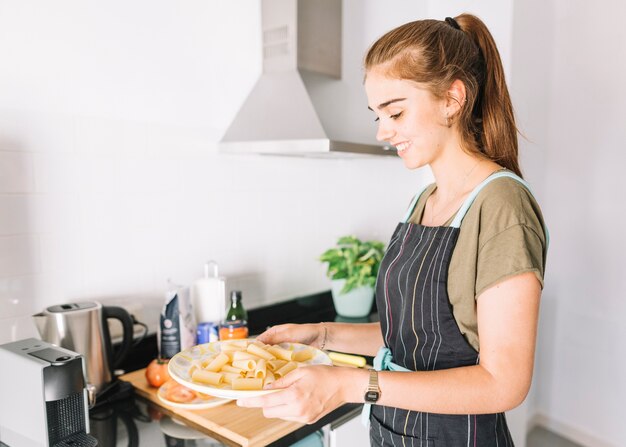 Jovem mulher de sorriso que prende o macarrão rigatoni cru na cozinha