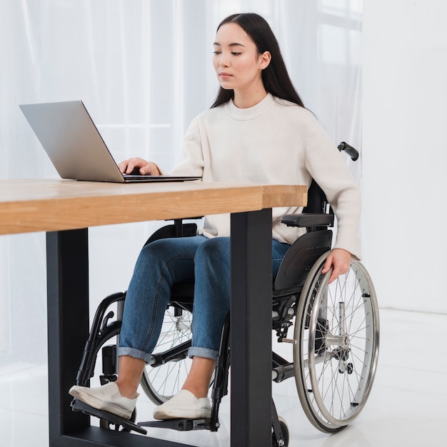 Jovem mulher deficiente que senta-se na cadeira de rodas usando o portátil