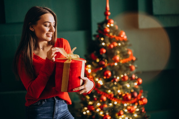 Jovem mulher desembalar presente de Natal pela árvore de Natal