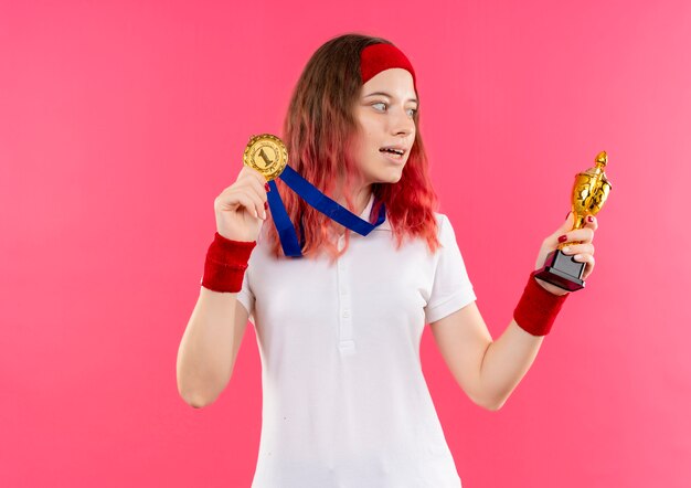 Jovem mulher desportiva com bandana e medalha de ouro pendurada no pescoço segurando troféu olhando para ele feliz e saiu de pé sobre a parede rosa