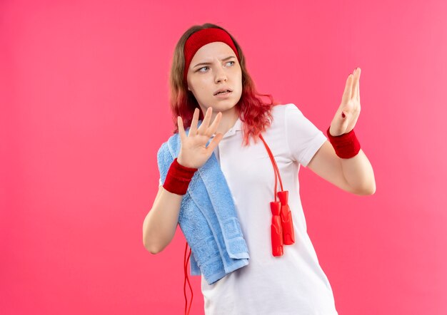 Jovem mulher desportiva com bandana e toalha no ombro fazendo gesto de defesa com as mãos olhando com expressão de medo em pé sobre a parede rosa