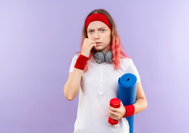 Jovem mulher desportiva com fones de ouvido segurando um tapete de ioga, parecendo confusa e preocupada em pé sobre a parede roxa