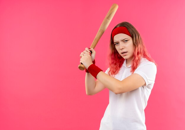 Jovem mulher desportiva com uma bandana jogando beisebol segurando um taco, parecendo confiante em pé sobre a parede rosa