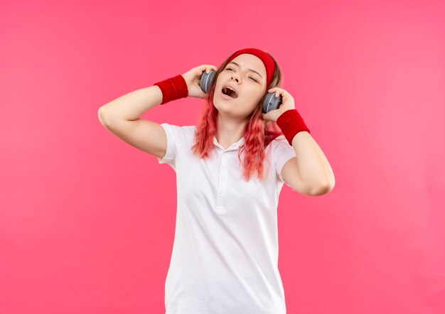 Jovem mulher desportiva com uma bandana na cabeça e fones de ouvido, curtindo sua música favorita e se sentindo feliz cantando uma música em pé sobre a parede rosa