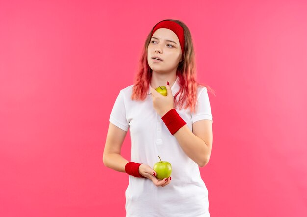Jovem mulher desportiva com uma bandana segurando duas maçãs, olhando para o lado com uma expressão pensativa em pé sobre a parede rosa