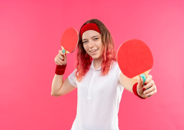 Jovem mulher desportiva com uma bandana segurando duas raquetes de tênis de mesa e sorrindo com uma cara feliz em pé sobre a parede rosa