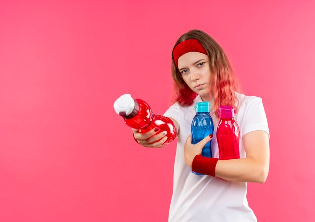 Jovem mulher desportiva com uma bandana segurando garrafas de água e uma das garrafas em pé sobre a parede rosa