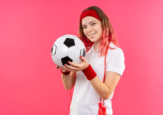 Jovem mulher desportiva com uma bandana segurando uma bola de futebol com um sorriso no rosto em pé sobre a parede rosa