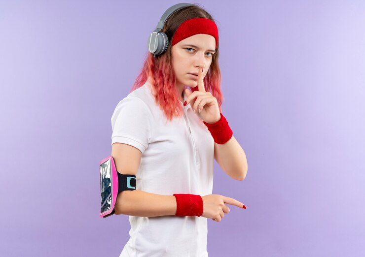 Foto grátis jovem mulher desportiva com uma faixa na cabeça e fones de ouvido fazendo gesto de silêncio com o dedo nos lábios em pé sobre a parede roxa