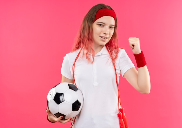 Jovem mulher desportiva com uma faixa na cabeça, segurando uma bola de futebol com o punho cerrado, feliz e saindo em pé sobre a parede rosa