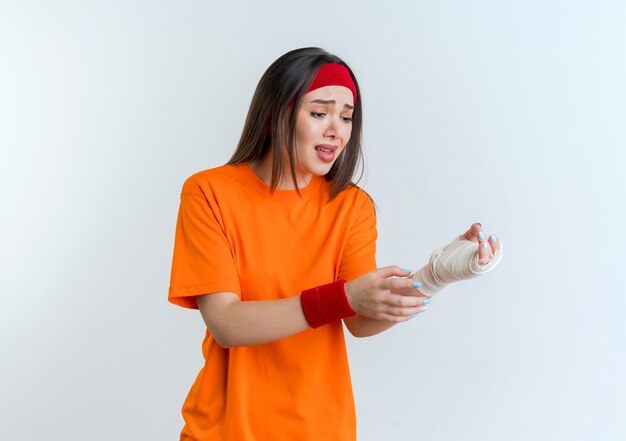 Jovem mulher desportiva e dolorida usando bandana e pulseiras, tocando e olhando para o pulso ferido envolto em bandagem isolada