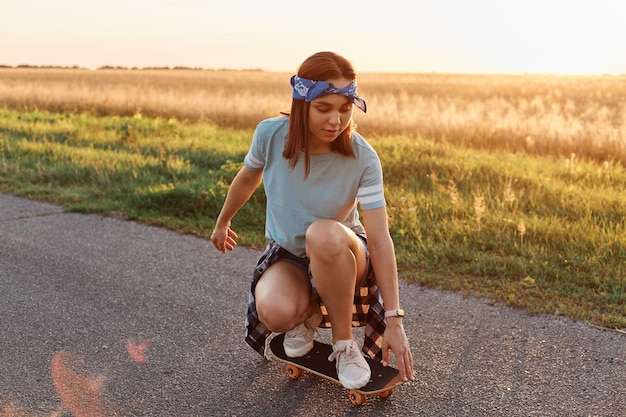 Jovem mulher desportiva vestindo camiseta e faixa de cabelo de cócoras no skate, andando de longboard na estrada de asfalto no verão, gastando o tempo do sol de forma ativa.