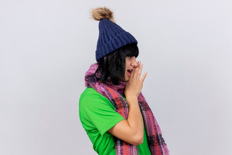 Foto grátis jovem mulher doente com chapéu de inverno e lenço em pé na vista de perfil, olhando em linha reta, mantendo a mão perto da boca, sussurrando, isolado na parede branca
