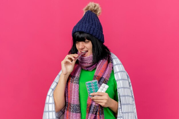 Jovem mulher doente usando chapéu de inverno e lenço embrulhado em manta segurando pacotes de comprimidos médicos e mordendo um deles, olhando para o lado isolado na parede rosa com espaço de cópia