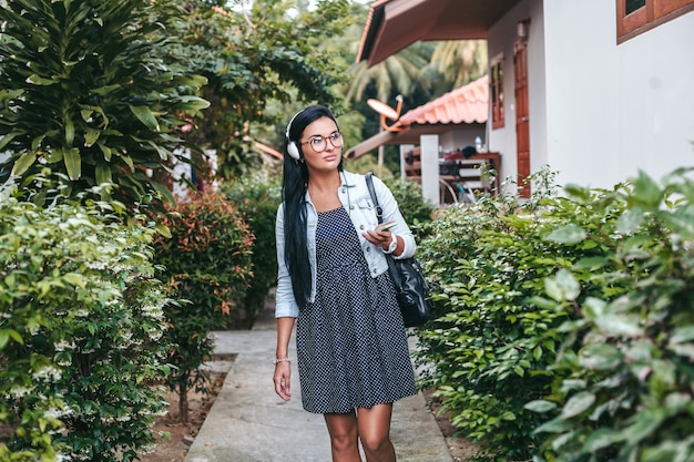 Jovem mulher elegante andando com smartphone, ouvindo música em fones de ouvido, férias de verão